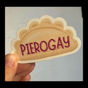 May include: A sticker featuring a cartoon pierogi with the word "PIEROGAY" in burgundy letters. The pierogi is light brown with a scalloped edge, and the background is a light blue.