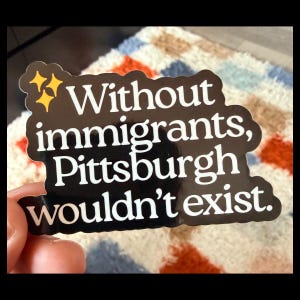 May include: Black sticker with white text that reads "Without immigrants, Pittsburgh wouldn't exist." The sticker has a black border and two yellow starbursts. The sticker is held by a person's fingers.