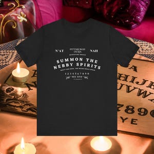 以下が含まれることがあります： 「SUMMON THE NEBBY SPIRITS」と「PITTSBURGH OUIJA」の白い文字がプリントされた黒いTシャツ。シャツには数字の1〜0と「SEE YINZ」の文字も表示されています。シャツはろうそくが立てられたテーブルの上に置かれています。