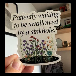 May include: A sticker with the text "Patiently waiting to be swallowed by a sinkhole." The sticker features a floral design with purple, red, and orange flowers. The sticker is held by a hand.