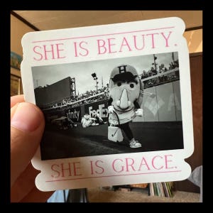 May include: A black and white photo of a mascot wearing a baseball uniform and holding a bucket. The photo is framed with a white border and the text "SHE IS BEAUTY. SHE IS GRACE." in pink letters.