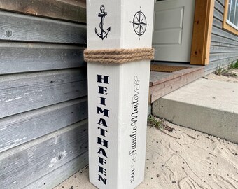 May include: A white wooden signpost with nautical motifs, including a black anchor and compass rose. The signpost features the word "HEIMATHAFEN" in black and is wrapped with rope. The text "PERSONALISIERBAR" is at the top.