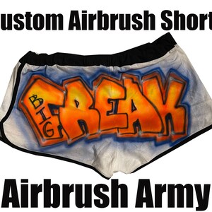 May include: White airbrush shorts with a black waistband and trim. The shorts feature orange graffiti-style lettering that spells out "FREAK" with the word "BIG" above it. The text "Custom Airbrush Shorts" and "Airbrush Army" are also visible.