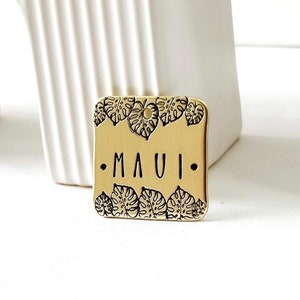 Könnte beinhalten: Ein quadratisches goldenes Schild mit einem tropischen Blattdesign um den Rand. Das Schild trägt die Aufschrift "MAUI" in schwarzen Buchstaben.