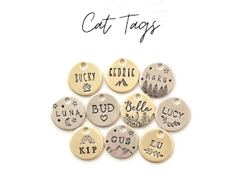 May include: A collection of ten round, gold, silver, and brass cat ID tags with various engraved designs and names, including "Bucky", "Cedric", "Haku", "Luna", "Bud", "Bella", "Lucy", "Kip", "Gus", and "Lu".