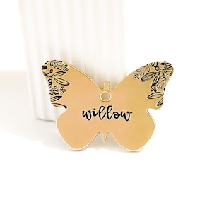 Op de afbeelding: Een gouden vlinder-vormige tag met een bloemenpatroon en het woord "willow" in zwart gedrukt.