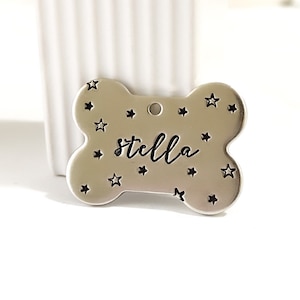 May include: Silver bone-shaped dog tag with a star pattern and the name "Stella" engraved in black.