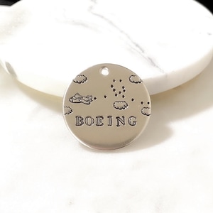 May include: Silver charm with a small airplane flying above clouds and the word "BOEING" engraved on the front.
