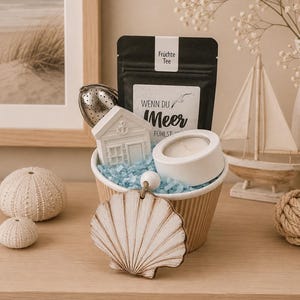Könnte beinhalten: Ein Geschenk-Set mit maritimem Thema, mit einem Muschelschmuck, einem kleinen weißen Haus, einer Kerze und einem Teebeutel. Der Korb enthält blaues Papier und befindet sich neben einem kleinen Segelboot und einem gerahmten Strandbild.