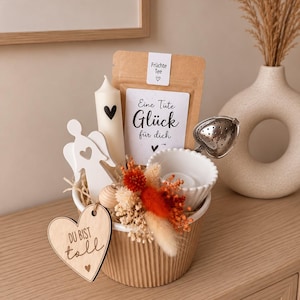 Könnte beinhalten: Ein Geschenkset in einem gerippten beigen Behälter mit einer braunen Papiertüte mit der Aufschrift "Früchte Tee" und "Eine Tüte Glück für dich", einer weißen Kerze mit Herz, einem weißen Engel, einem herzförmigen Tee-Ei und einem Holzherz-Anhänger mit der Aufschrift "DU BIST toll".