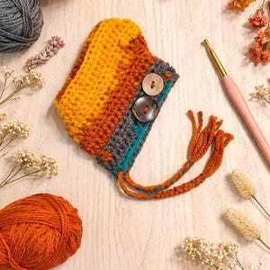 Könnte beinhalten: Ein handgefertigtes Häkelstück in Gelb-, Orange- und Grautönen mit zwei Holzknöpfen und orangefarbenen Quasten. Das Bild zeigt Wollknäuel, Trockenblumen und eine Häkelnadel, was auf ein Bastelprojekt hindeutet.