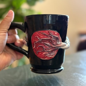 May include: Black ceramic mug with a unique, textured red and white design on the side. The mug has a black handle and a glossy finish. The design appears to be a stylized anatomical form.