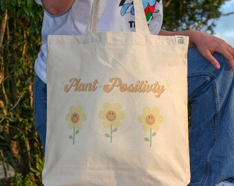 May include: A white canvas tote bag with the text "Plant Positivity" in yellow script. Three yellow flowers with smiley faces are printed below the text.