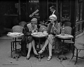 May include: Two women in 1920s fashion sit at outdoor cafe tables in Paris, France. The women are wearing hats and dresses. They are drinking coffee and talking. The cafe tables are round and have white tablecloths. The cafe chairs are wicker and have black cushions. The cafe is located on a street corner and has a sign that says "Telephone".