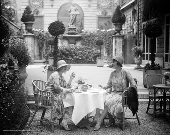 May include: Two people wearing hats sit at a table outside in a garden setting. They are having tea and a conversation. The table is set with a white tablecloth and teacups. There are plants and trees in the background.