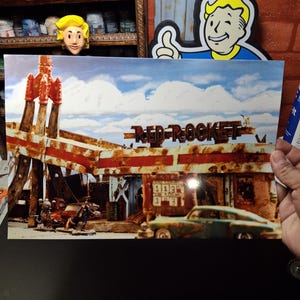 May include: A print of the Red Rocket gas station from the Fallout video game series. The image shows a rusty, dilapidated building with a classic car in front. The sky is blue with white clouds, and the text "Red Rocket" is visible.