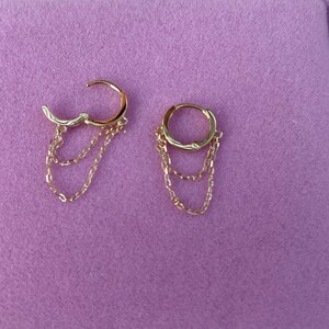 May include: A pair of gold hoop earrings with three dangling chains. The hoops are simple and the chains are delicate.