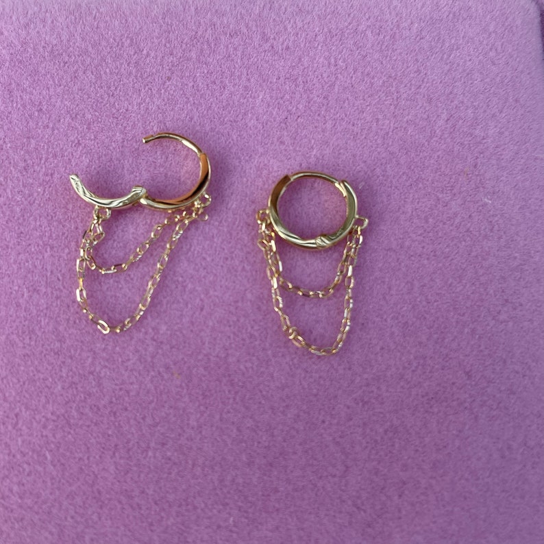 May include: A pair of gold hoop earrings with three dangling chains. The hoops are simple and the chains are delicate.