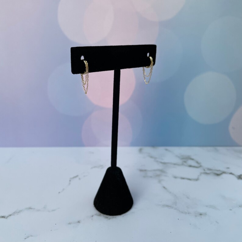 May include: A pair of gold hoop earrings with a dangling chain. The earrings are displayed on a black stand.