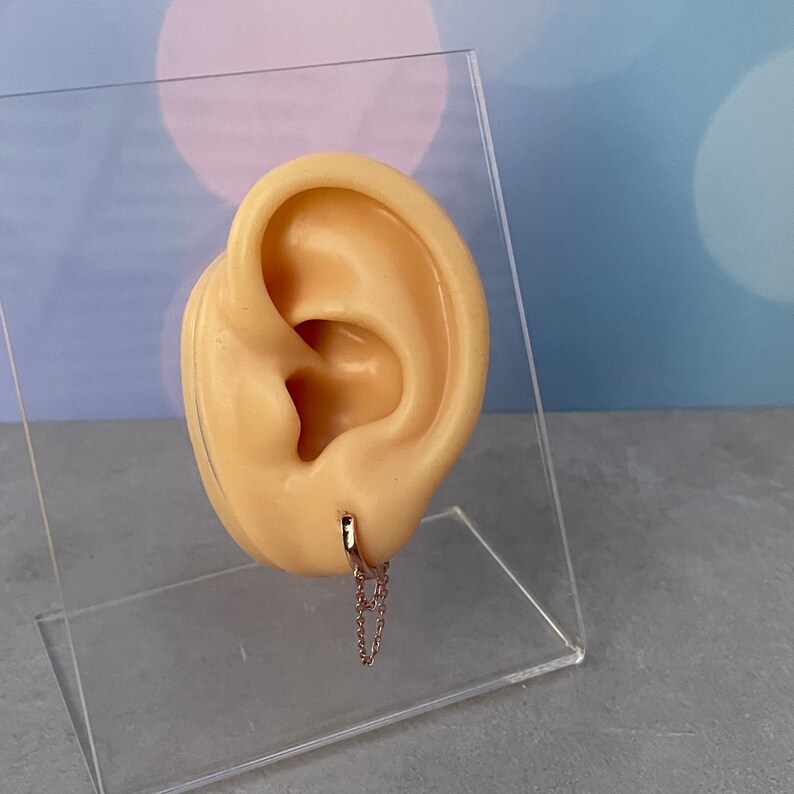 May include: A gold hoop earring with a delicate chain dangling from it. The earring is shown on a model ear.