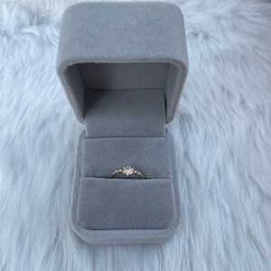 May include: A delicate gold ring with a small cluster of clear stones set in the center, displayed in a gray velvet ring box.
