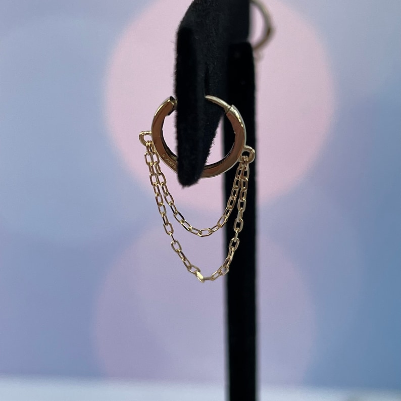 May include: Gold hoop earrings with a delicate chain dangling from the bottom. The chain is made of two strands of small, linked circles.