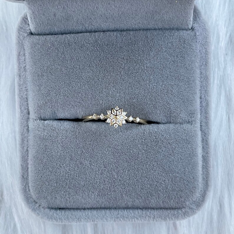May include: A delicate gold ring with a snowflake design set with small diamonds. The ring is displayed in a gray velvet ring box.