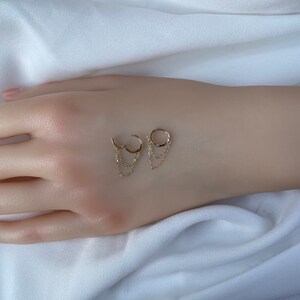 May include: A pair of gold hoop earrings with dangling chains. The earrings are on a white surface.