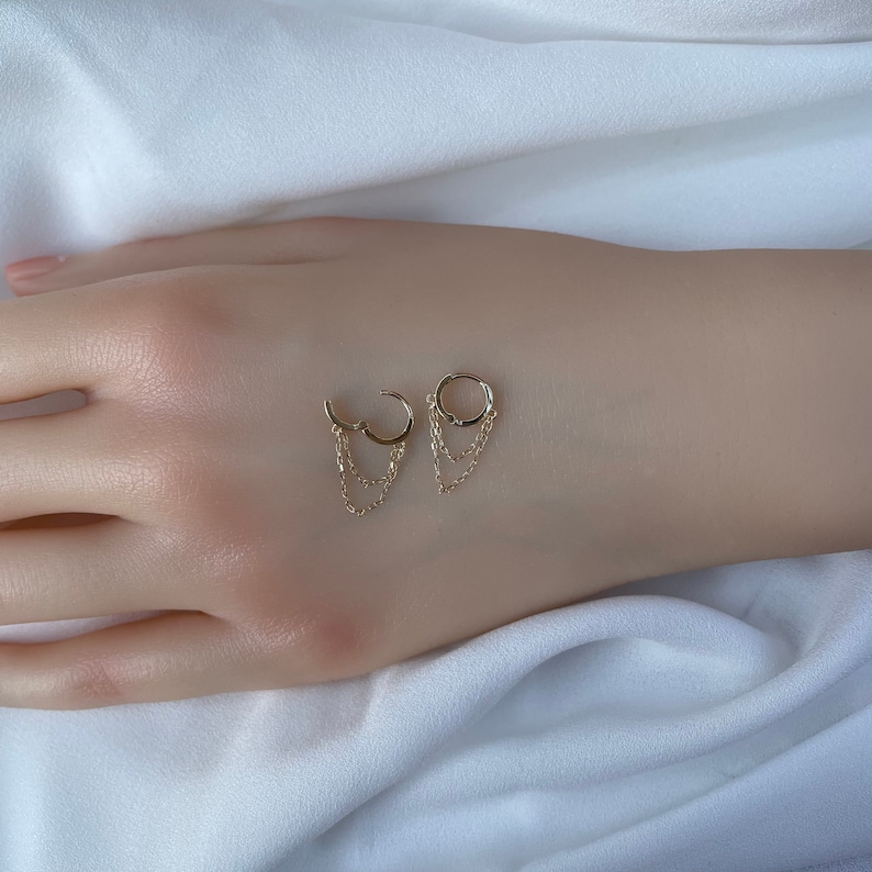 May include: A pair of gold hoop earrings with dangling chains. The earrings are on a white surface.