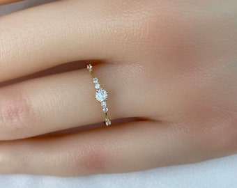 May include: A delicate gold ring featuring a central round-cut diamond, accented by smaller diamonds along the band. The ring is set on a finger, showcasing its elegant design and sparkle. The ring's band is thin and minimalist.