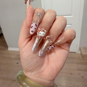 May include: Long stiletto-shaped press-on nails featuring a mix of clear, glittery gold, and light pink finishes. The design includes leopard print patterns, small clear rhinestones, and a three-dimensional Hello Kitty character charm.