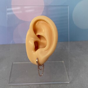 May include: A gold hoop earring with a delicate chain dangling from it. The earring is displayed on a mannequin ear.