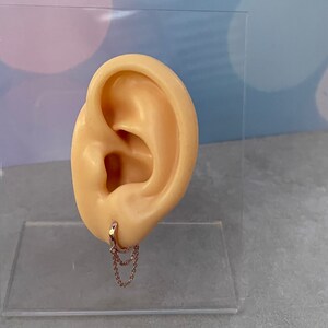 May include: A close-up of a fake ear with a rose gold hoop earring with a double chain dangling from it.