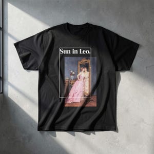 Puede incluir: Camiseta negra con una pintura vintage enmarcada de dos mujeres, una con un vestido rosa, reflejadas en un espejo. El texto "Sun in Leo." est&aacute; encima de la pintura. La camiseta es de material suave.