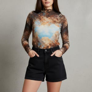 Op de afbeelding: Een doorschijnende top met lange mouwen en een wolkenprint in bruin, blauw en wit. Gecombineerd met een zwarte spijkerbroekshort. De top heeft een hoge, gerimpelde halslijn en gerimpelde manchetten.