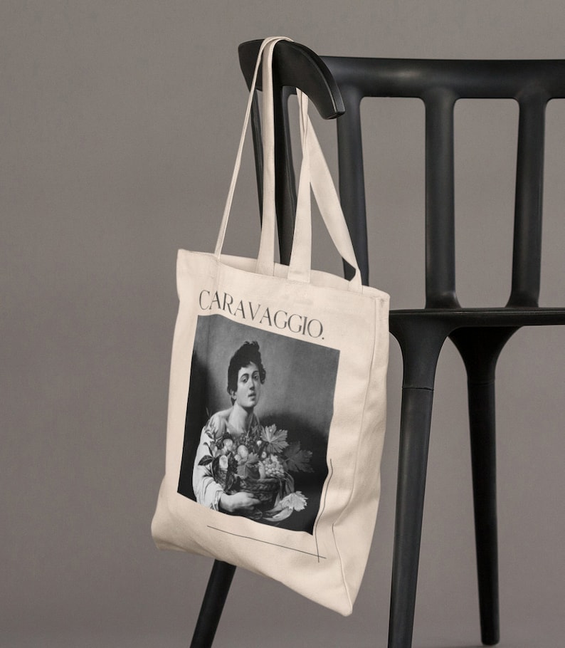 May include: A beige canvas tote bag with black handles and a black and white image of a man holding a basket of fruit. The text "CARAVAGGIO." is printed on the bag.