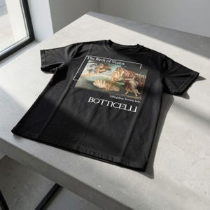 May include: Black t-shirt featuring Botticelli's "The Birth of Venus" artwork. The design includes the painting's title and artist's name in white text. The shirt is made of a soft material and is displayed on a light gray surface.