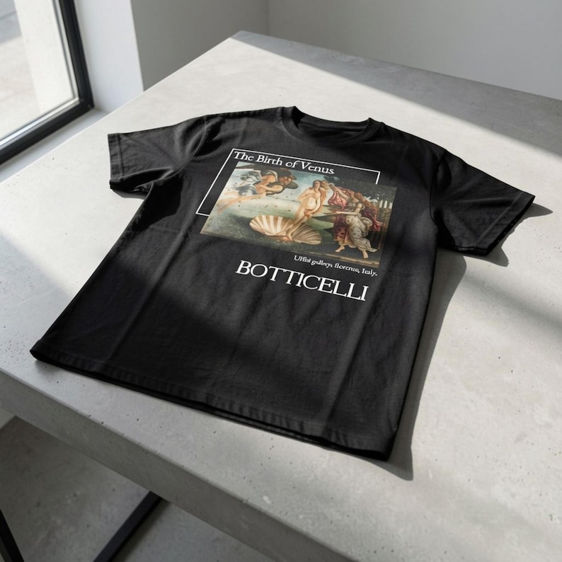 May include: Black t-shirt featuring Botticelli's "The Birth of Venus" artwork. The design includes the painting's title and artist's name in white text. The shirt is made of a soft material and is displayed on a light gray surface.