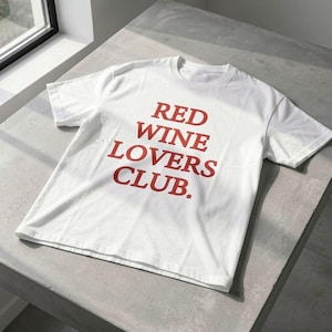 Può includere: T-shirt bianco con la scritta "RED WINE LOVERS CLUB" in rosso. La maglietta è realizzata in un materiale morbido e viene esposta su una superficie grigia, con una finestra sullo sfondo.