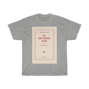 Op de afbeelding: Een lichtgrijs t-shirt met een boekcover-ontwerp. Het ontwerp toont de titel "LE DEUXI&Egrave;ME SEXE" in het rood, met de naam van de auteur en het logo van de uitgever. Het t-shirt is gemaakt van een zacht, comfortabel materiaal.