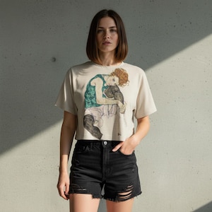 Könnte beinhalten: Beiges Crop-Top mit einem Aufdruck einer sitzenden Figur mit roten Haaren, die ein grünes Oberteil und weiße Shorts trägt. Das Model trägt auch schwarze Jeansshorts mit Distressed-Details.
