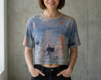 Puede incluir: Una camiseta corta con una pintura de escena acuática en tonos azules, naranjas y grises. La camiseta de manga corta tiene cuello redondo y un punto rojo en la parte superior derecha. La camiseta se combina con pantalones cortos vaqueros negros.