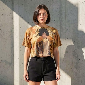 Puede incluir: Una camiseta de manga corta y cuello redondo con un estampado gráfico en tonos dorados y marrones de un retrato con un sombrero negro grande. Se combina con unos pantalones cortos negros lisos de talle alto con cierre de botón y cremallera.