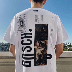 Puede incluir: Camiseta blanca con un estampado gráfico en blanco y negro de una pintura de Hieronymus Bosch. El texto "BOSCH." está impreso verticalmente en el lado izquierdo de la camiseta. El texto "Dark side of the forest The shutter on the right represents Hell It describes a dream world demanic oppressive and of innumerable torments" está impreso en el lado derecho de la camiseta.