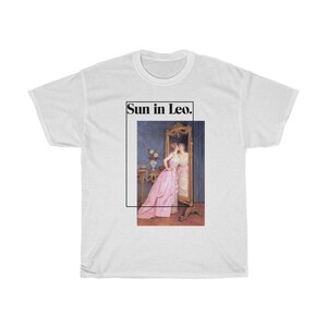 Puede incluir: Camiseta blanca con una imagen enmarcada de dos mujeres, una con un vestido rosa, reflejadas en un espejo. El texto "Sun in Leo." est&aacute; encima de la imagen. La camiseta es de estilo unisex.