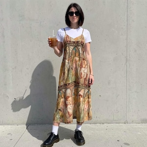 May include: A midi-length slip dress featuring a repeating stained-glass style print with figures in muted earth tones, worn over a plain white short-sleeved t-shirt. The dress has thin spaghetti straps and a v-neckline, paired with black lace-up shoes.