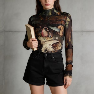 Puede incluir: Una camiseta transparente de manga larga con un estampado detallado de una pintura histórica, combinada con unos shorts vaqueros negros. La camiseta tiene un cuello alto y la obra de arte cubre toda la prenda. Un libro se sostiene en la mano del modelo.