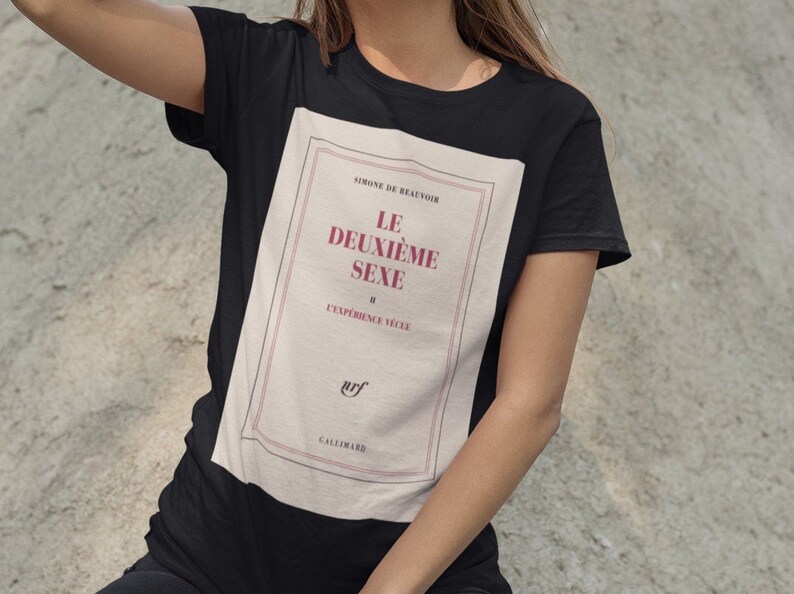 Op de afbeelding: Zwart T-shirt met een boekcover ontwerp. Het ontwerp bevat de titel "LE DEUXI&Egrave;ME SEXE" in bordeauxrode tekst, de naam van de auteur en het logo van de uitgever. Het T-shirt heeft een klassieke ronde hals.