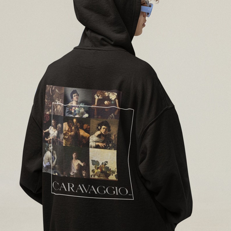 Pode incluir: Camisola de moletom com capuz preta com uma estampa gr&aacute;fica de uma colagem de pinturas de Caravaggio. O texto "CARAVAGGIO." &eacute; impresso abaixo da colagem.