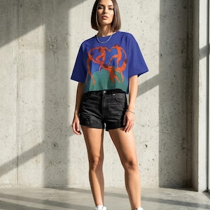 May include: A blue crop top with an orange illustration of dancing figures. The model is wearing black denim shorts and white high-top trainers. The background is a concrete wall.